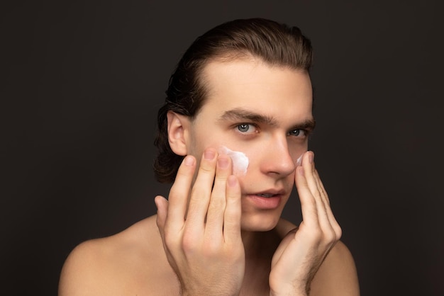Ritratto di giovane uomo bello che applica crema per il viso prendendosi cura della pelle isolata su sfondo marrone scuro