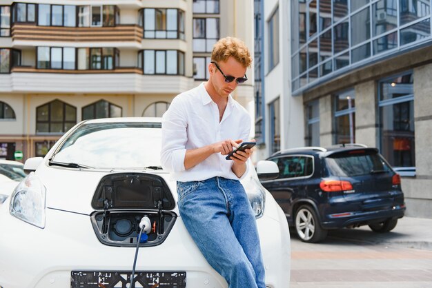 充電ステーションに立って、カジュアルな服装で若いハンサムな男の肖像画。エコ電気自動車のコンセプト