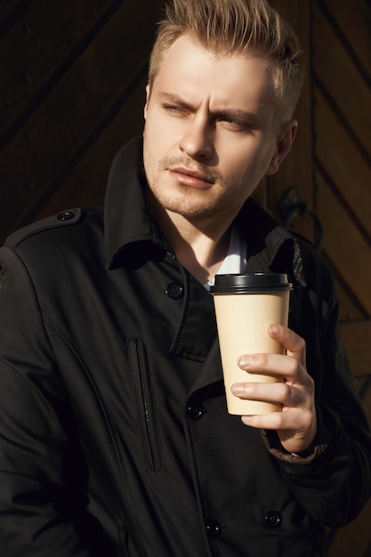 Portrait of young handsome man in dark coat with cup of coffee standing near old gate