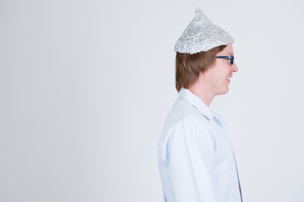portrait of young handsome man doctor wearing tin foil hat as conspiracy theory concept on white