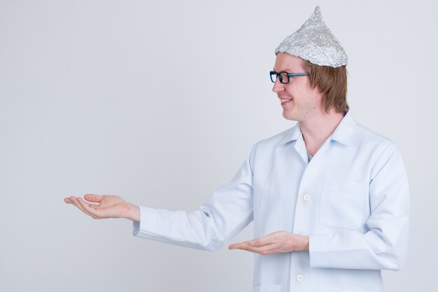Portrait of young handsome man doctor wearing tin foil hat as conspiracy theory concept on white