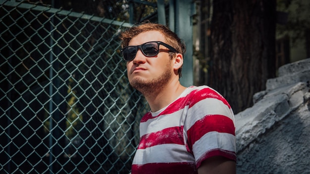 Photo a portrait of a young handsome man in a tshirt and in sunglasses near a lattice fence in the park