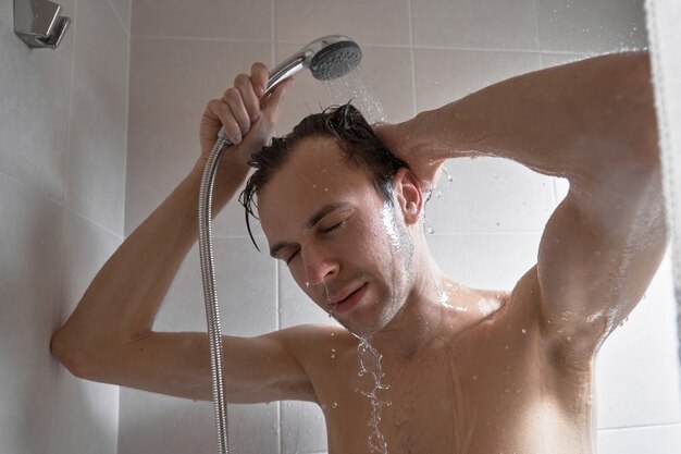 Portrait of young handsome man washes himself with shower gel, lathers head with shampoo in the bathroom at home close-up