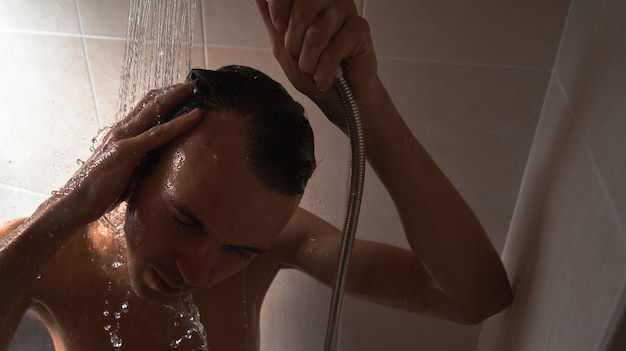 Portrait of young handsome man washes himself with shower gel, lathers head with shampoo in the bathroom at home close-up