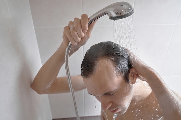 Portrait of young handsome man washes himself with shower gel, lathers head with shampoo in the bathroom at home close-up