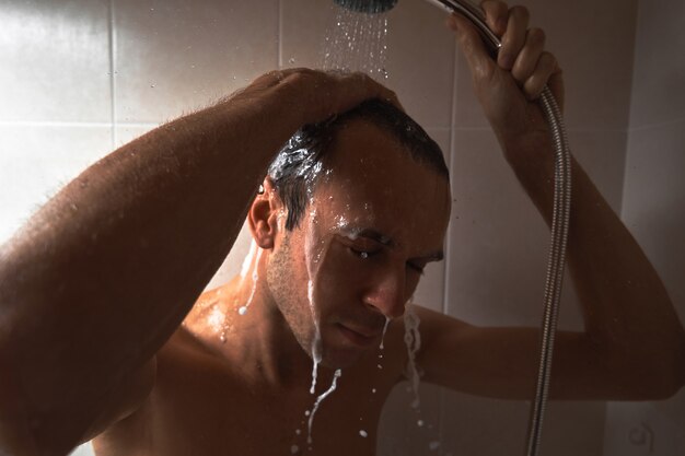 Portrait of young handsome man washes himself with shower gel, lathers head with shampoo in the bathroom at home close-up