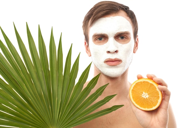 Portrait of young handsome man with cosmetic mask on his face holding orange in hand isolated on exotic palm leaves background