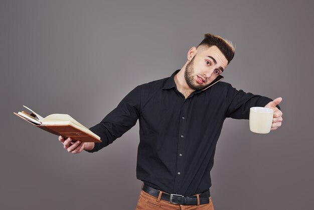 Portrait of young handsome man with phone, cup of coffe and book on hands.