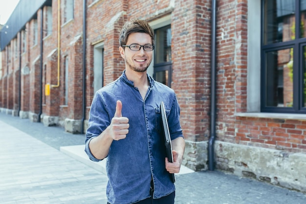 Portrait of a young handsome student freelancer with glasses Walks on the streets of the city holds a laptop in his hands points a finger super looks at the camera smiles