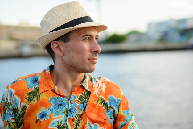 Portrait of young handsome tourist man at pier on view of the river in Bangkok city