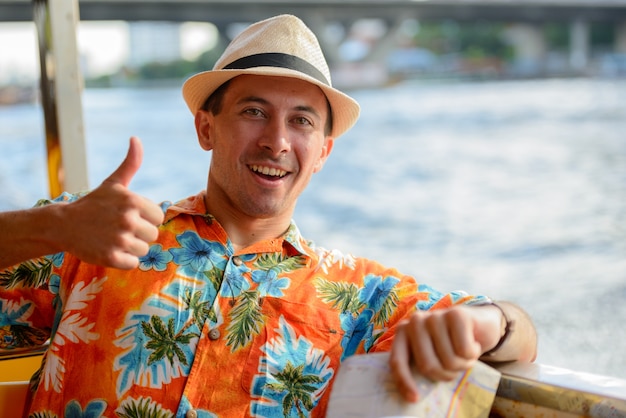 portrait of young handsome tourist man riding boat as public transportation in Bangkok city