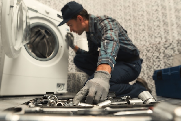 Photo portrait of young handyman in uniform sitting next to broken washing machine and trying to fix it. horizontal shot. side view. selective focus on a hand and toolbox