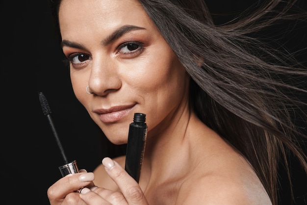 Portrait of a young happy beautiful naked woman posing isolated over black wall holding mascara apply makeup.