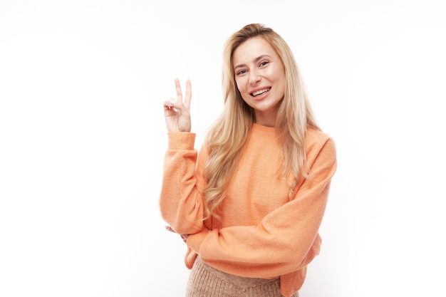 Portrait of young happy blonde woman in orange sweater showing peace and love gesture with fingers isolated on white studio background