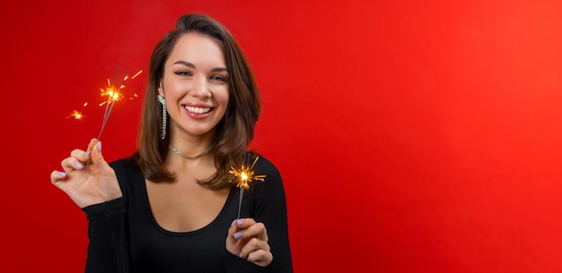 Portrait young happy brunette woman in black blouse holding with sparklers on red background with copy space