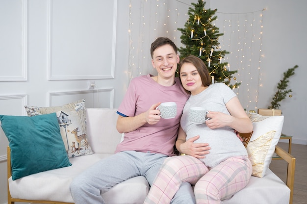 Portrait of a young happy couple relaxing at home with mugs in their hands. Christmas time and baby waiting.