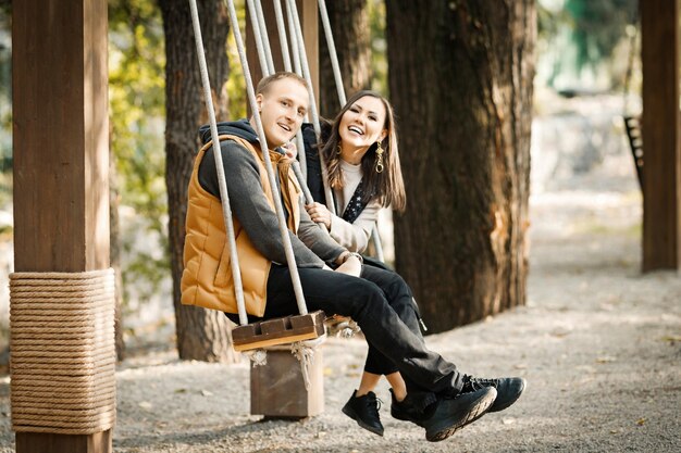 Portrait of young happy couple riding on a swing in autumn in warm clothes in basic colors in a park outdoors