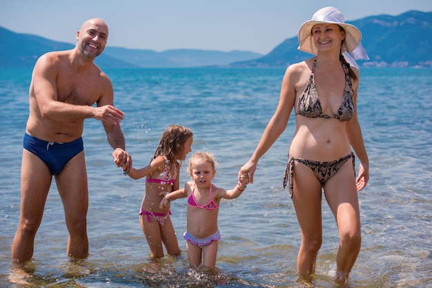 портрет молодой счастливой пары с милыми маленькими дочерьми, весело проводящими время, стоя на мелководье моря во время летних каникул Концепция здорового семейного отдыха