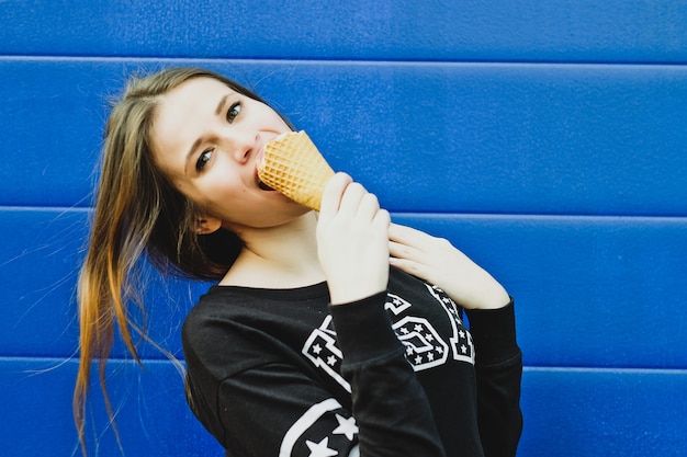 Portrait of young happy girl eating ice-cream, outdoor, over blue wall background