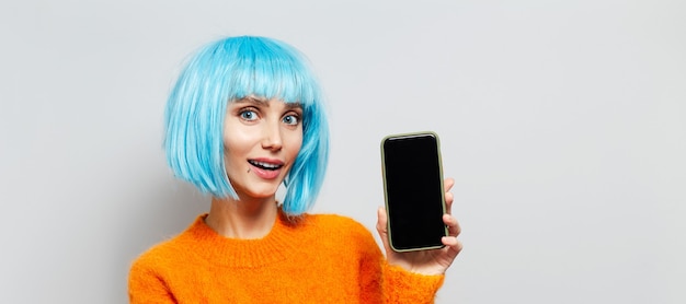 Portrait of young happy girl holding smartphone in hand, wearing blue hair wig and orange sweater on white background with copy space. Panoramic banner view.