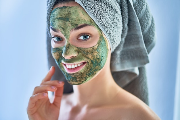 Ritratto di giovane femmina in buona salute felice in asciugamano di bagno con la maschera dell'argilla del fronte dopo la doccia durante il giorno della stazione termale a casa. cura della pelle del viso