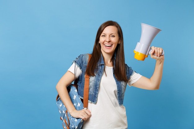 Portrait of young happy joyful attractive woman student in denim clothes with backpack holding electronic megaphone isolated on blue background. Education in university. Copy space for advertisement.