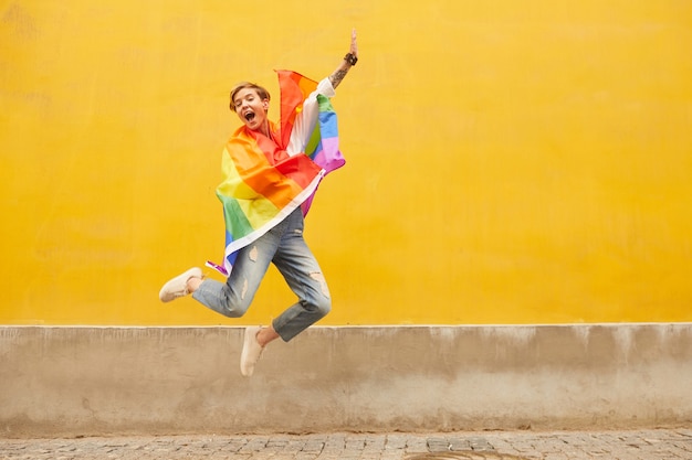 Портрет молодой счастливой лесбиянки с цветным флагом на плечах, прыгающей на открытом воздухе