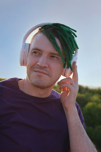 Portrait of young happy man with green dreadlocks wireless headphones listening to music in nature in sunny weather in evening