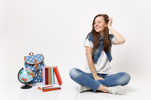 Portrait of young happy pretty woman student with closed eyes correcting hairstyle sitting near globe, backpack, school books