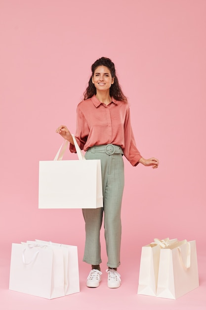 Photo portrait of young happy shopaholic standing among shopping bags and smiling against the pink background