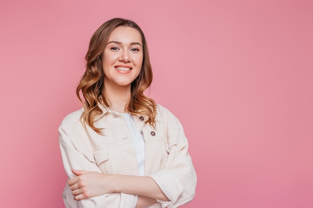 Photo portrait of a young happy smiling caucasian student girl with wavy hair in a beige corduroy jacket isolated on pink wall  girl standing with her hands folded