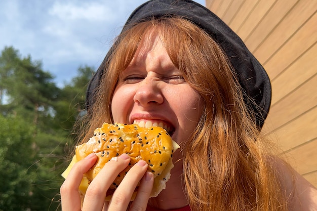 Portrait of young happy woman is eating biting juicy delicious burger outside cafe looking at camera