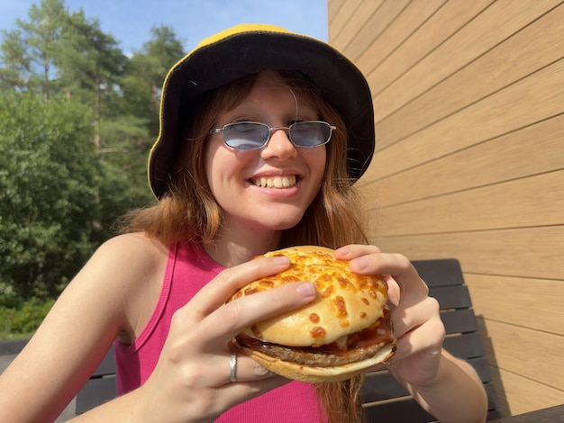 Portrait of young happy woman is eating biting juicy delicious burger outside cafe looking at camera