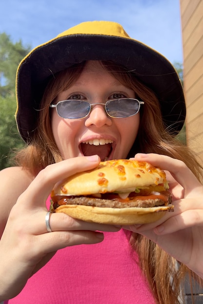 Photo portrait of young happy woman is eating biting juicy delicious burger outside cafe looking at camera