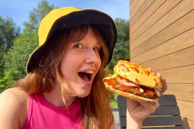 Foto il ritratto di giovane donna felice sta mangiando l'hamburger mordace e delizioso fuori dal caffè che guarda l'obbiettivo