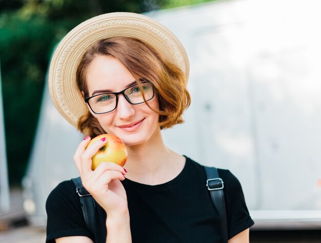 Portrait of young hipster woman in glasses and hat holding apple and smiling at the camera outdoors