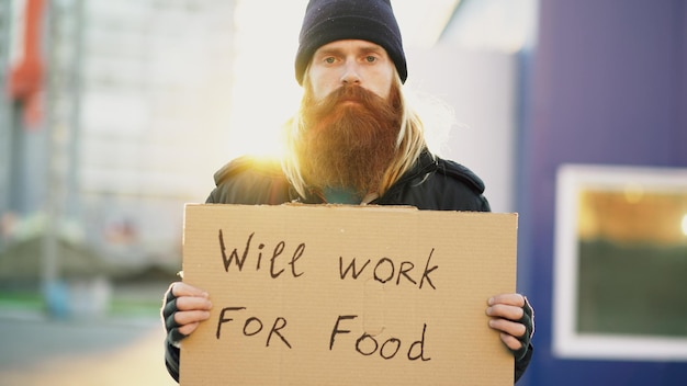 Portrait of young homeless man with cardboard looking at camera and wants to work for food