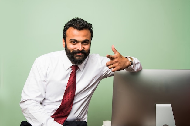 Portrait of young Indian businessman in beard, sitting in relaxed position at at office table with pleasant look. Confident asian male business person