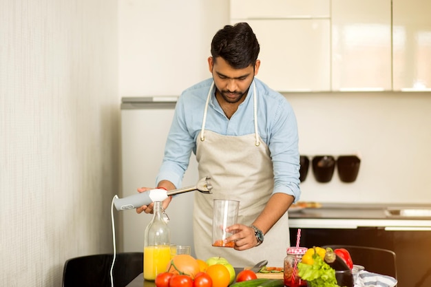 Portrait of young indian handsome man wearing apron standing in kitchen, holding electric blender, looking at camera and prepearing fresh juice or smoothie. Food, healthy meal concept.