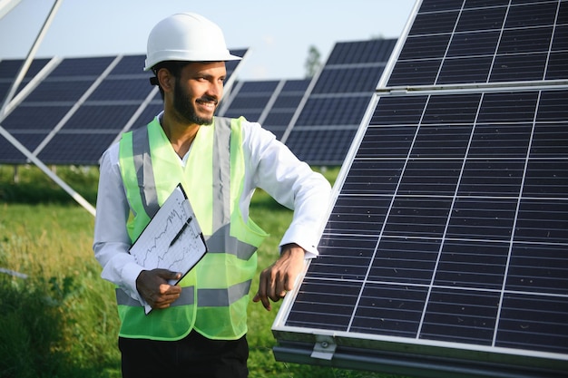 Portrait of Young indian man technician wearing white hard hat standing near solar panels against blue skyIndustrial worker solar system installation renewable green energy generation concept