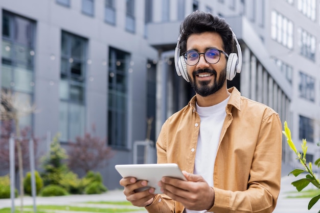 Portrait young indian man with headphones and tablet watching online video sitting on bench near