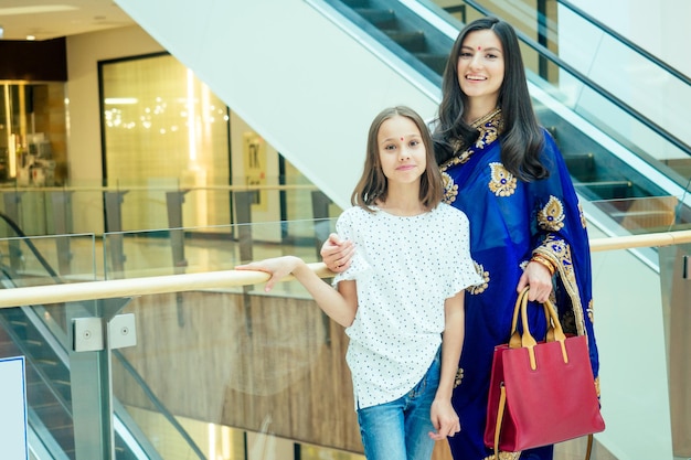 Portrait of a young indian woman wearing blue sari and gold\
bracelet having fun with her cute daughter in shopping\
mall,escalator background.red religious dot on forehead.