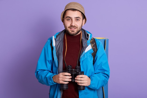 Portrait of young intrepid explorer wearing brown panama, red sweatshirt, blue jacket, holding binoculars, having sleeping pad and bag aback, posing isolated over lilac background. Travelling concept