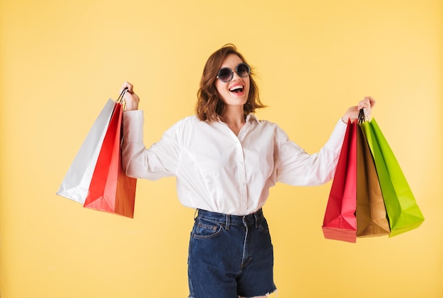 Portrait of young joyful lady in sunglasses standing with colorful shopping bags in hands on over pink background Happy woman standing in white shirt and denim shorts