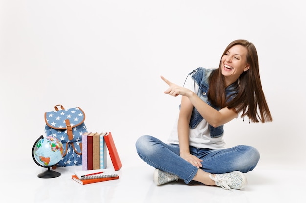 Portrait of young joyful laughing woman student pointing index finger aside sitting near globe backpack, school books isolated on white wall