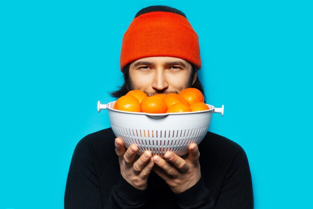 Photo portrait of young joyful man, with orange beanie hat, holding a bowl of mandarins. on the wall of blue wall.