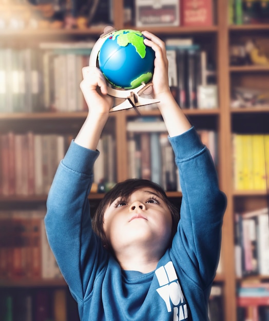 Ragazzino del ritratto che tiene globo in alto sopra la sua testa che osserva in su da sotto con la faccia curiosa, ragazzo del bambino che impara circa la geografia, l'istruzione e il concetto di homeschooling