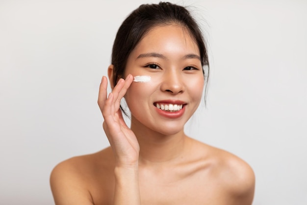 Portrait of young korean lady applying facial cream under her eyes taking care of skin standing over