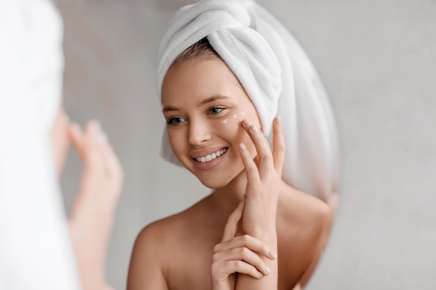 Foto ritratto di giovane donna che applica una crema per gli occhi nutriente dopo la doccia guardandosi allo specchio e sorridendo