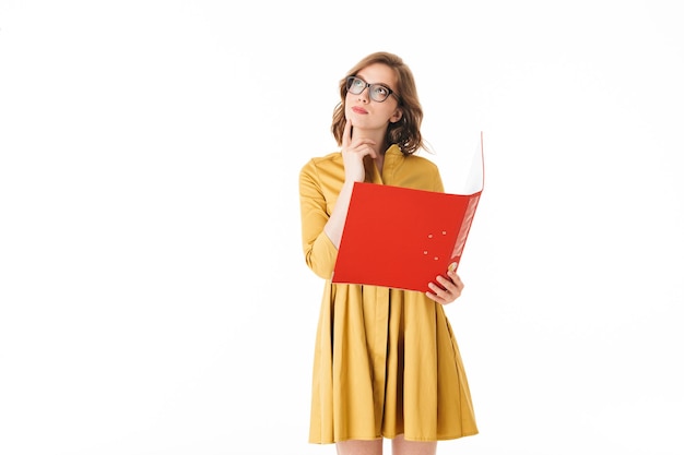 Portrait of young lady in eyeglasses and yellow dress standing with red folder in hand and dreamily looking aside on white background isolated